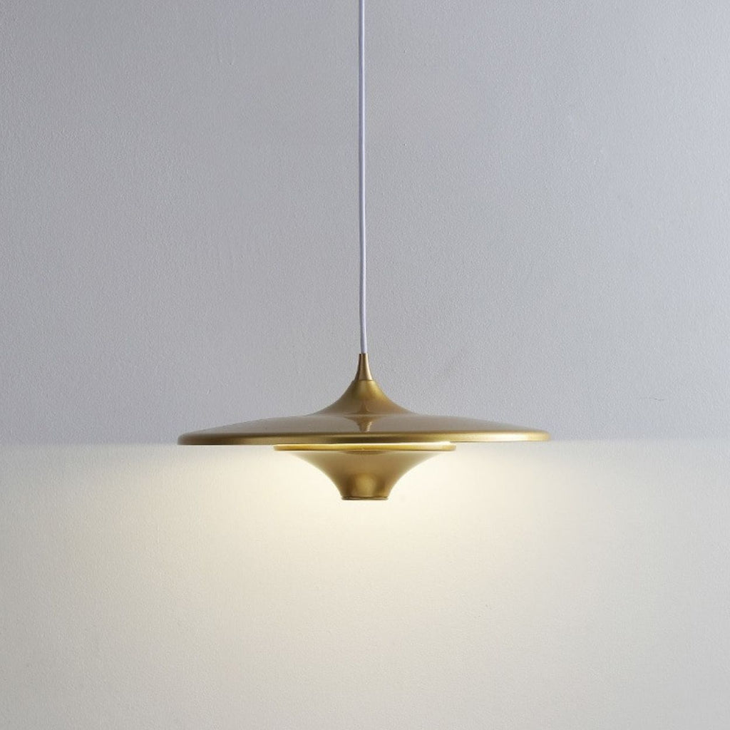 A modern, gold pendant light with a wide, flat shade hangs from a white cord against a plain, light-colored wall, casting a soft, even glow below it.