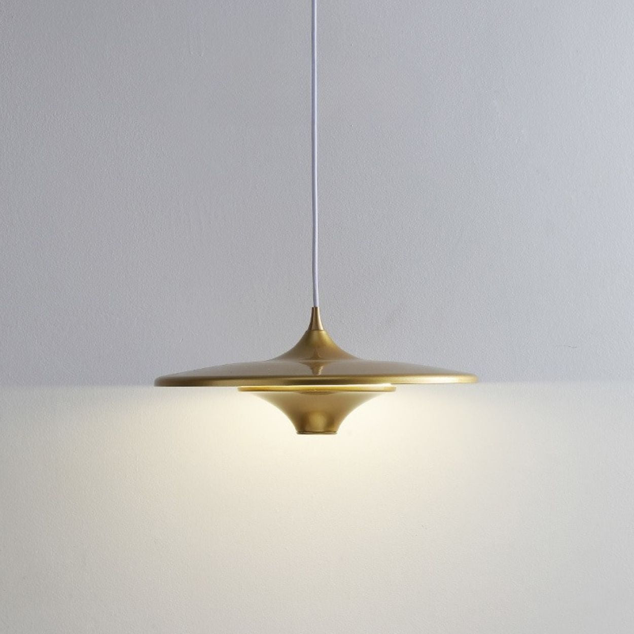 A modern, gold pendant light with a wide, flat shade hangs from a white cord against a plain, light-colored wall, casting a soft, even glow below it.