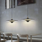 Two modern pendant lights hang above a simple table with three light-colored chairs against a gray wall in a minimalistic dining area.