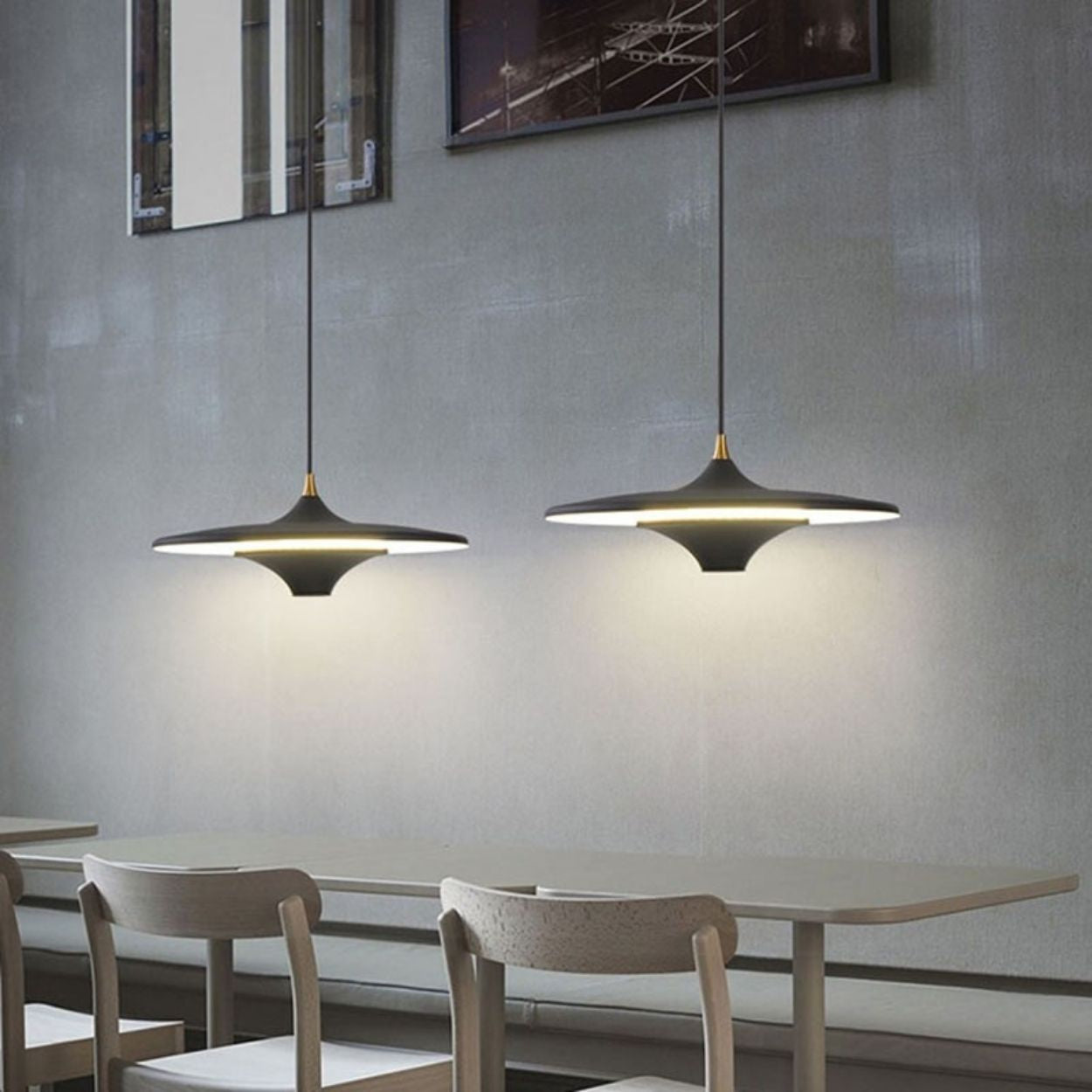 Two modern pendant lights hang above a simple table with three light-colored chairs against a gray wall in a minimalistic dining area.