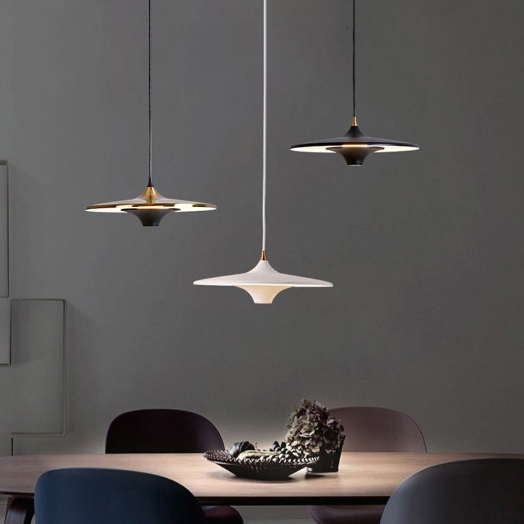 Three modern pendant lights—two dark, one white—hang above a wooden dining table with four chairs. A decorative bowl with pinecones sits at the center of the table, set against a minimalist gray background.