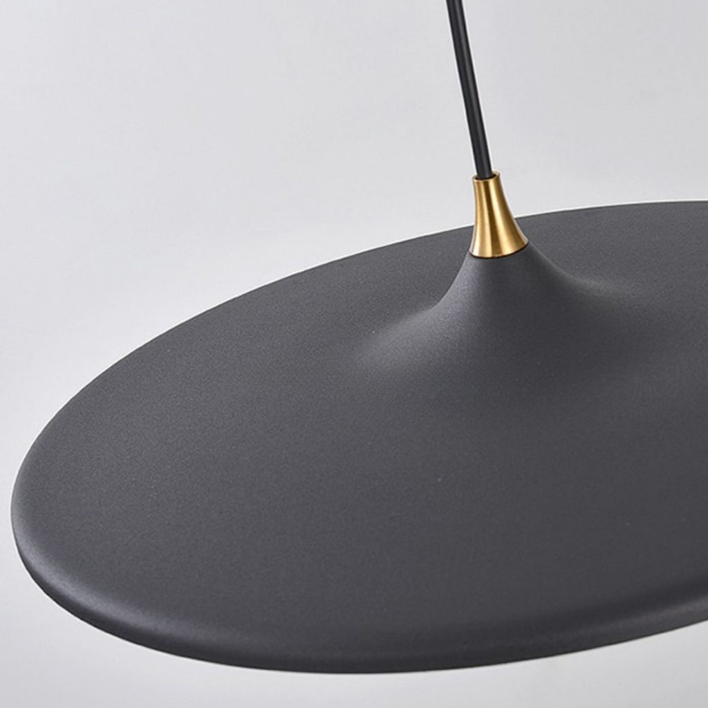 A close-up of a modern pendant light features a smooth, matte black shade and a gold accent near the top, hanging from a black cord against a plain light background.