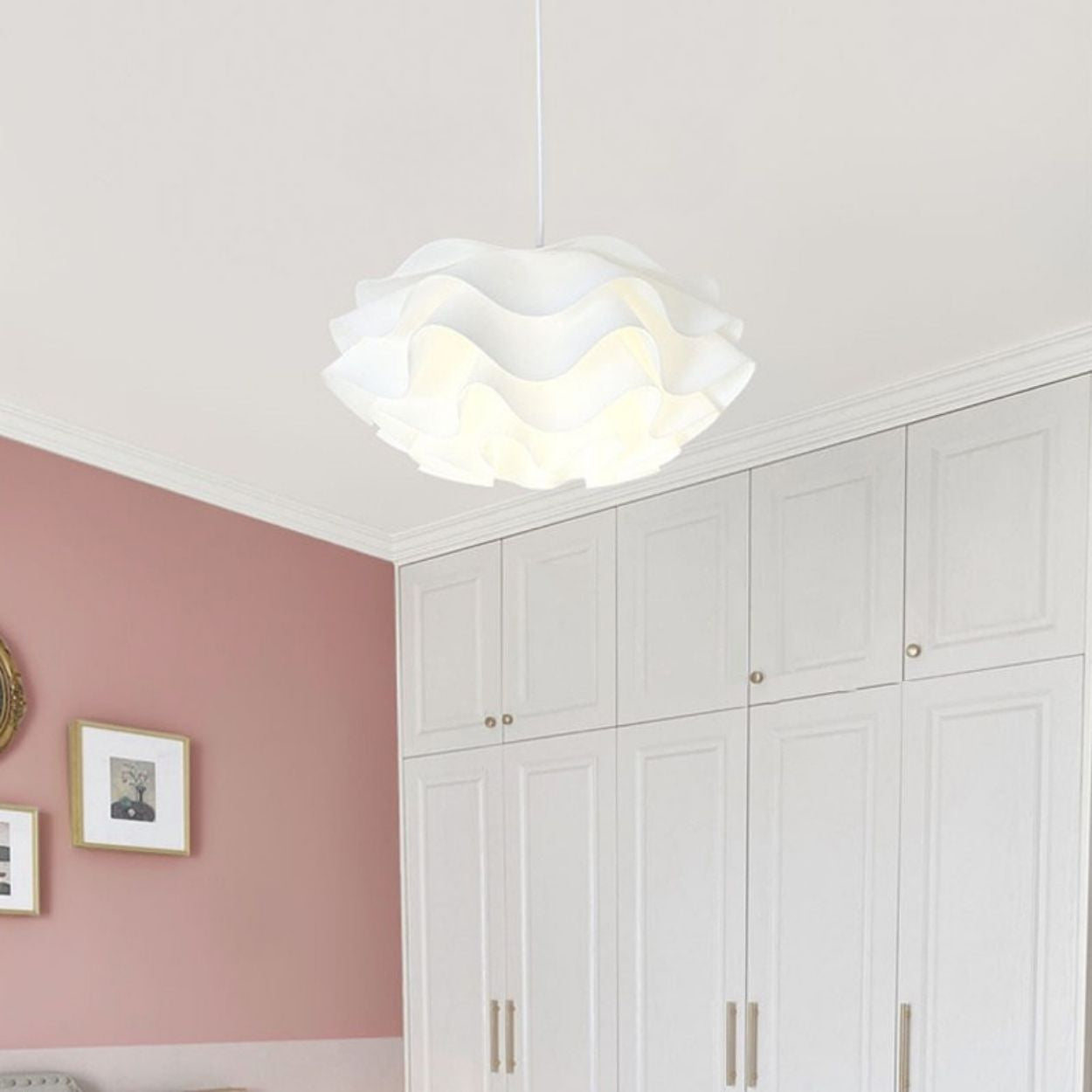 A modern white pendant light with a wavy design hangs from the ceiling in a room featuring white cabinets, pink walls, and framed artwork.