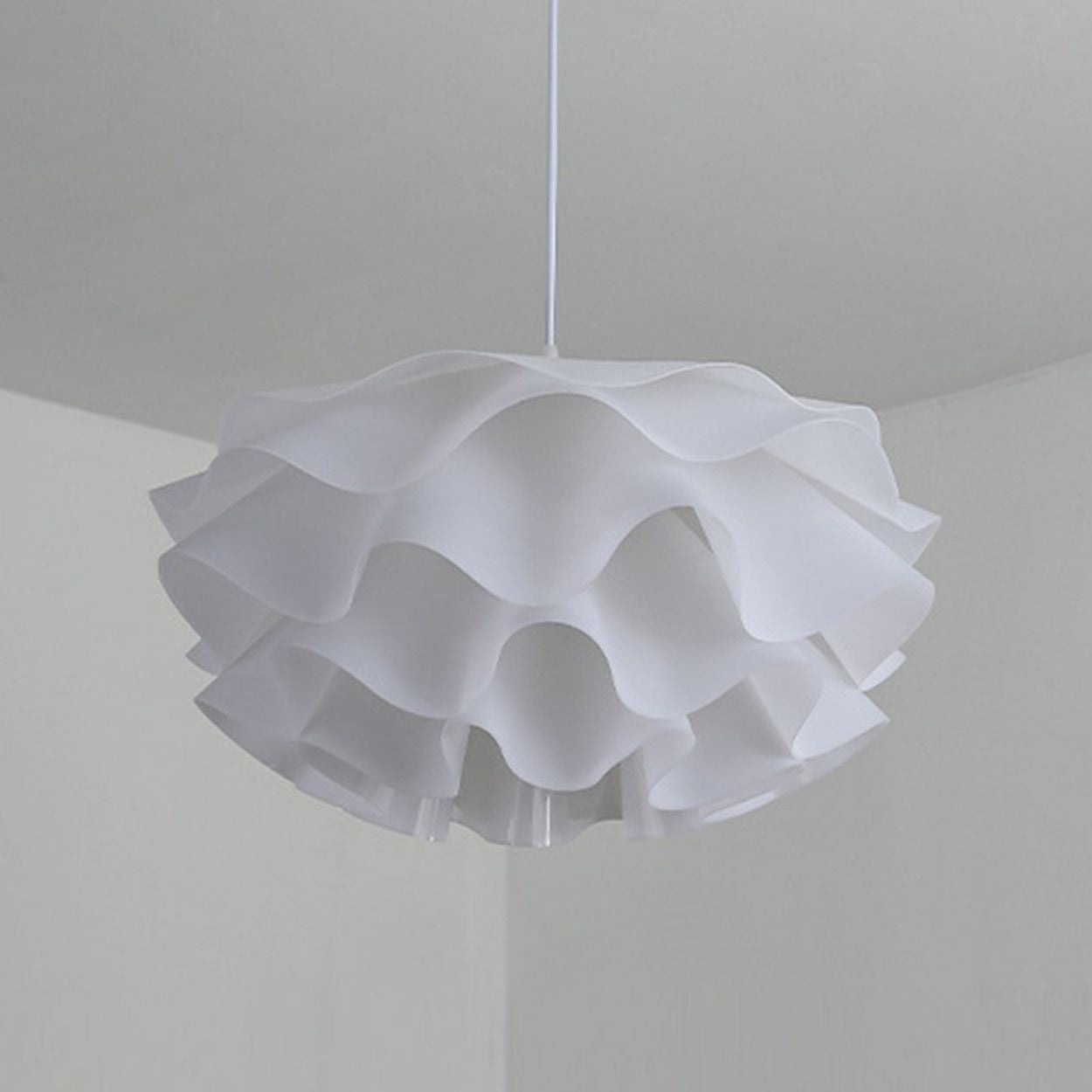 A modern pendant light with a wavy, ruffled white lampshade hangs from the ceiling in a minimalist room with light-colored walls.