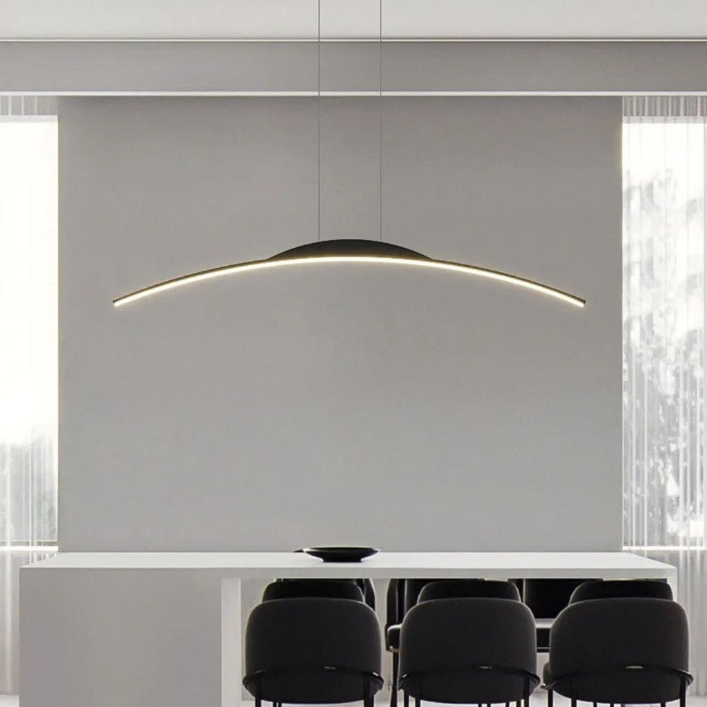 A modern dining room features a long white table, six black chairs, and a striking pendant light hanging above. Large windows with sheer curtains flood natural light into the minimalist space.
