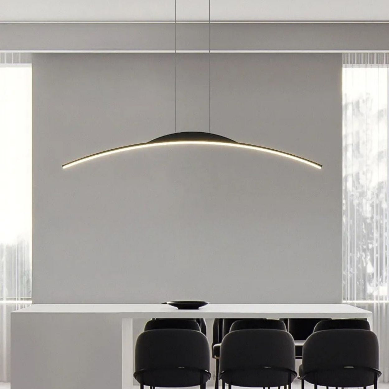 A modern dining room features a long white table, six black chairs, and a striking pendant light hanging above. Large windows with sheer curtains flood natural light into the minimalist space.