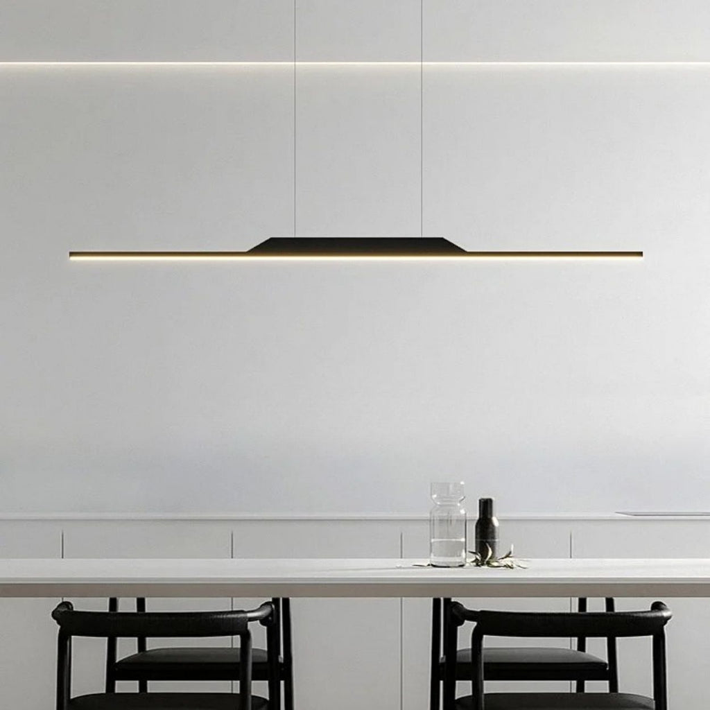 Minimalist dining area featuring a long, slim pendant light illuminating a white table, with two black chairs and a glass water carafe and cup set. The modern pendant light stands out against the plain white wall background.