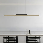 Minimalist dining area featuring a long, slim pendant light illuminating a white table, with two black chairs and a glass water carafe and cup set. The modern pendant light stands out against the plain white wall background.