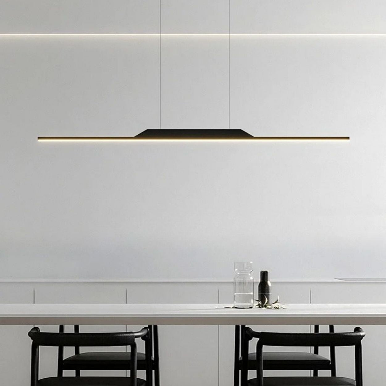 Minimalist dining area featuring a long, slim pendant light illuminating a white table, with two black chairs and a glass water carafe and cup set. The modern pendant light stands out against the plain white wall background.