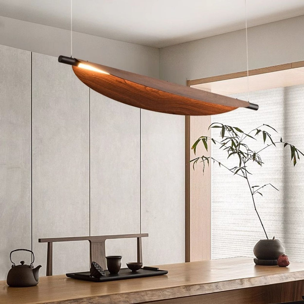 A modern dining area with a wooden table set for tea, a minimalist wood and metal chandelier overhead, and a simple potted plant against light-colored paneled walls.