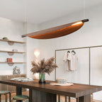 Modern dining area with a rectangular wooden table, a vase of pampas grass, wall shelves with bags and decor, and a unique curved wood chandelier pendant light hanging from the ceiling. Neutral tones and minimal decor throughout.