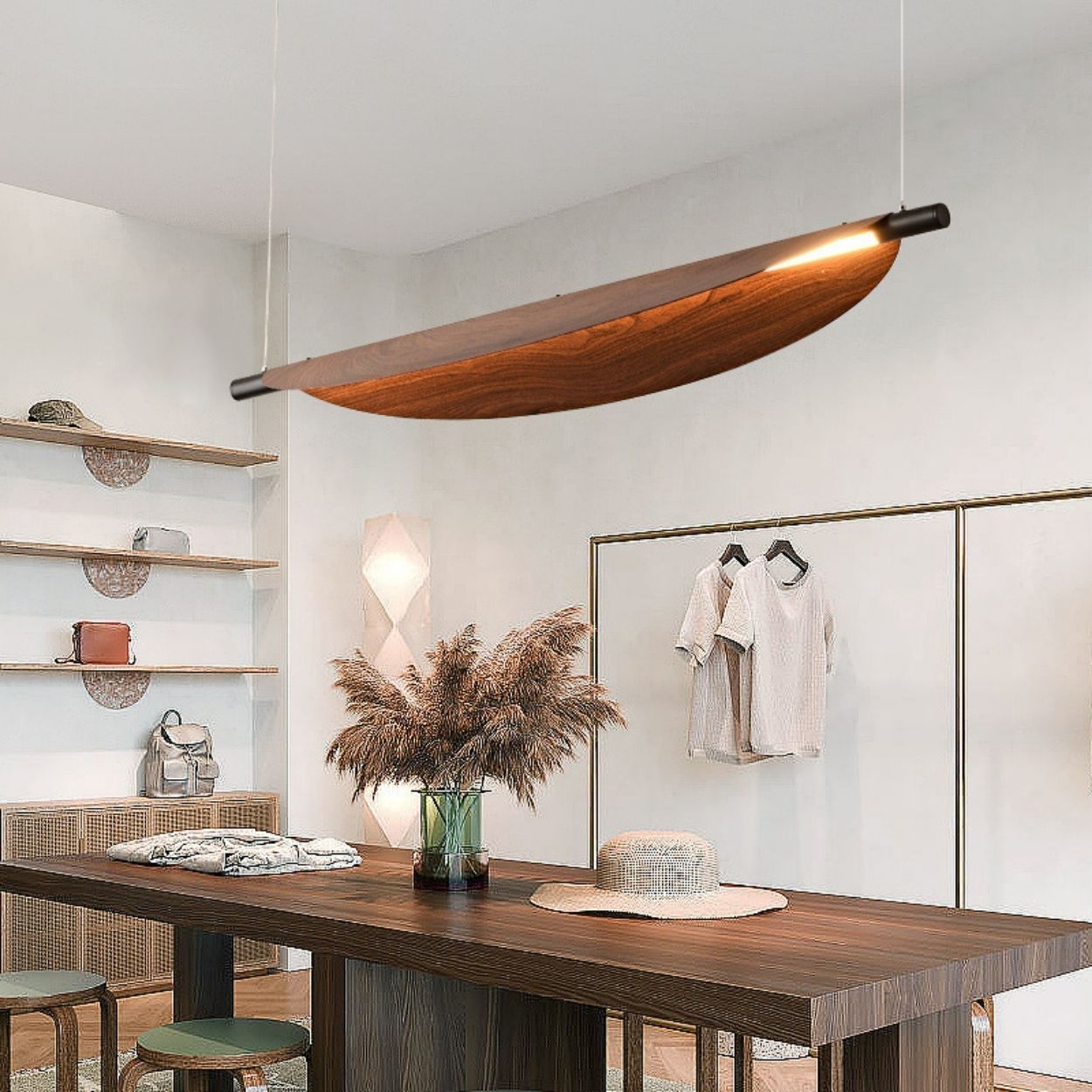 Modern dining area with a rectangular wooden table, a vase of pampas grass, wall shelves with bags and decor, and a unique curved wood chandelier pendant light hanging from the ceiling. Neutral tones and minimal decor throughout.