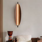 A modern wall light with an oval wooden frame and a central vertical light strip is mounted above a white cushioned chair. A decorative drum sits on a wooden table nearby, complementing the minimalist style of the wall and decor.