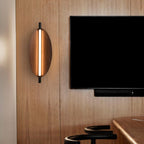 A modern wall sconce, or wall light, with a wooden oval design and vertical light strip is mounted on a wood-paneled wall next to a flat-screen TV. Part of a bar stool and wooden table are visible in the foreground.