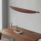 A minimalist dining area with a wooden table, two candlesticks with gray candles, and a modern, curved wooden chandelier suspended above against a plain gray wall.