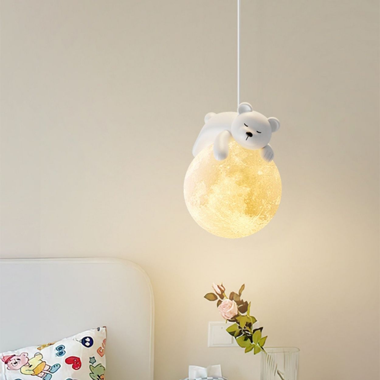 A cute pendant light shaped like a glowing moon with a small white bear figurine lying on top hangs above a bedside table with a vase and flower, next to a bed adorned with a colorful pillow.