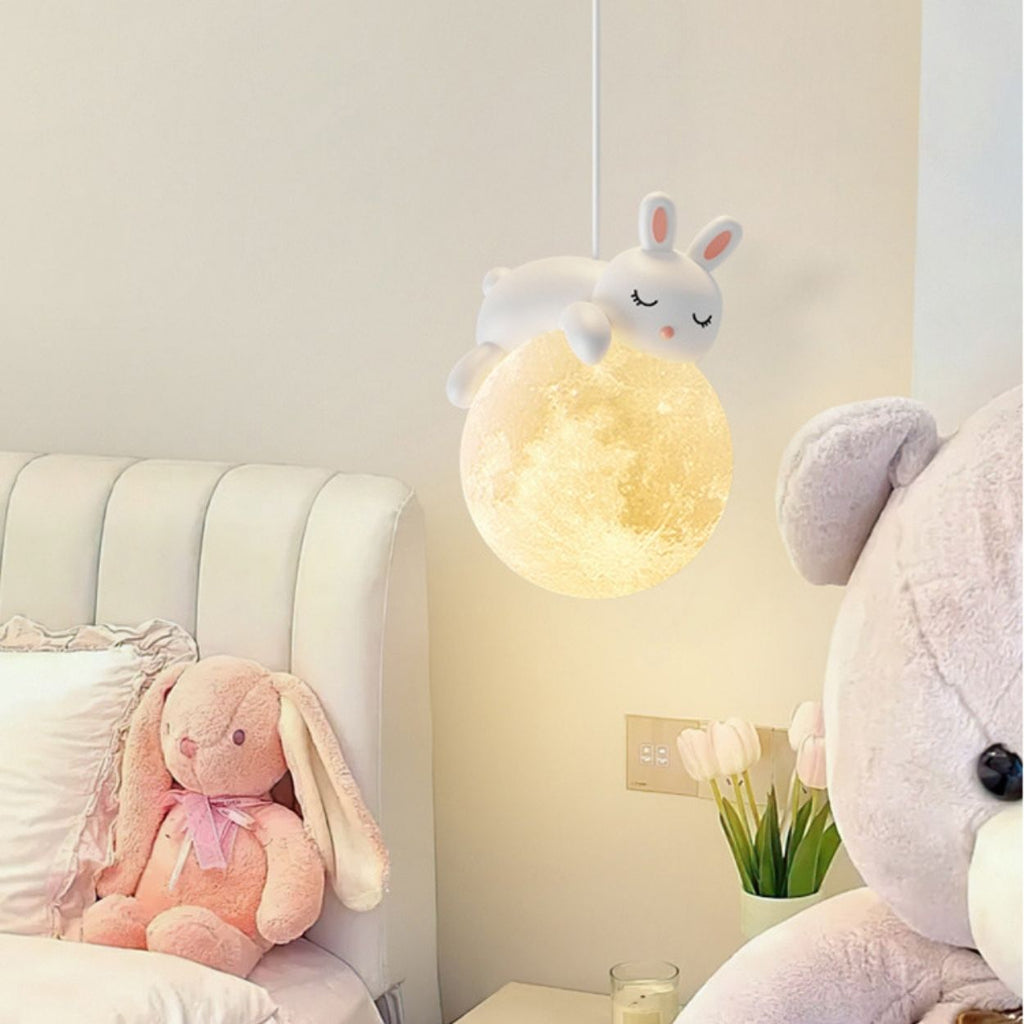A cozy bedroom with a plush white headboard, a pink stuffed bunny on the bed, a large teddy bear beside it, and a glowing moon-shaped pendant light featuring a sleeping bunny hanging beside a vase of tulips.