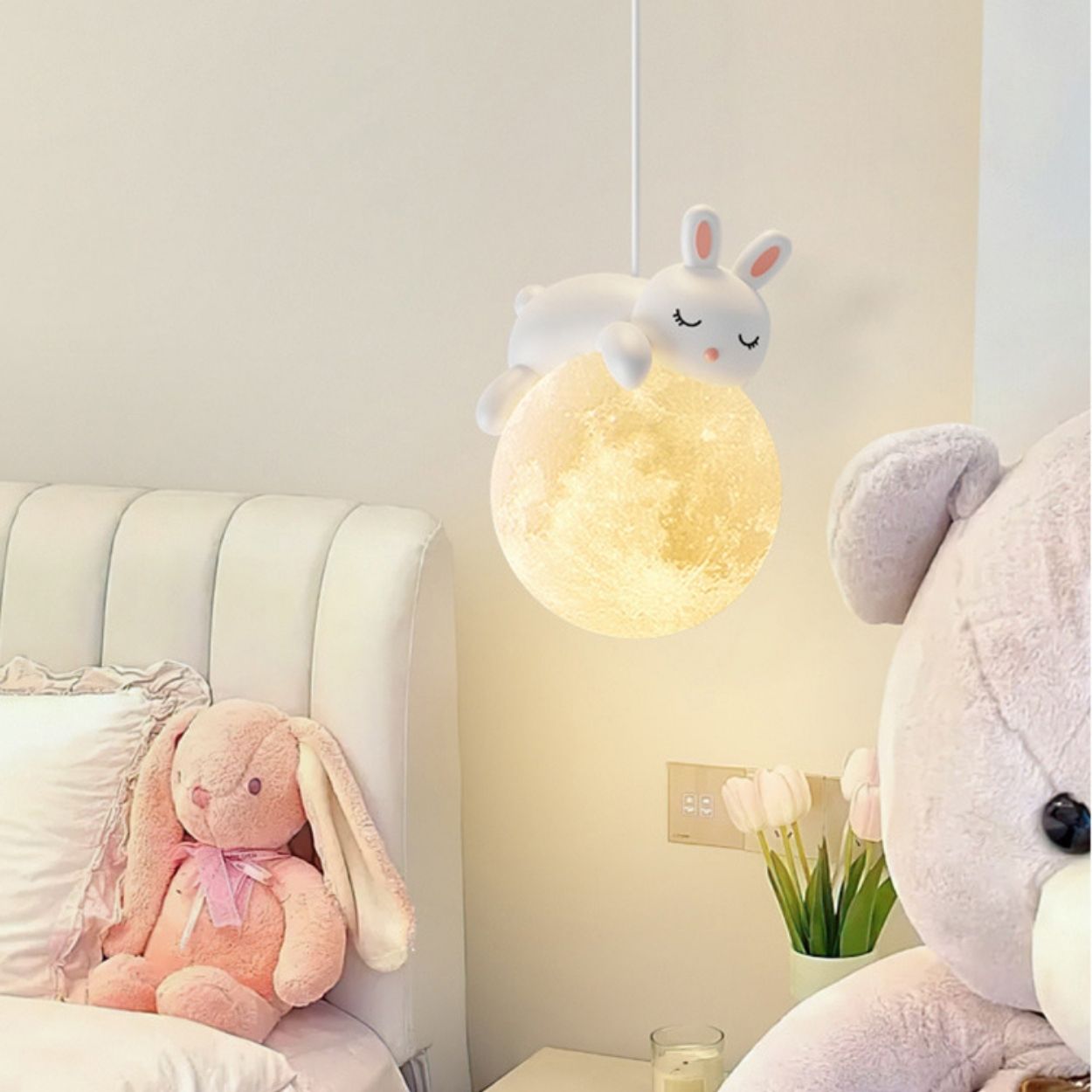 A cozy bedroom with a plush white headboard, a pink stuffed bunny on the bed, a large teddy bear beside it, and a glowing moon-shaped pendant light featuring a sleeping bunny hanging beside a vase of tulips.
