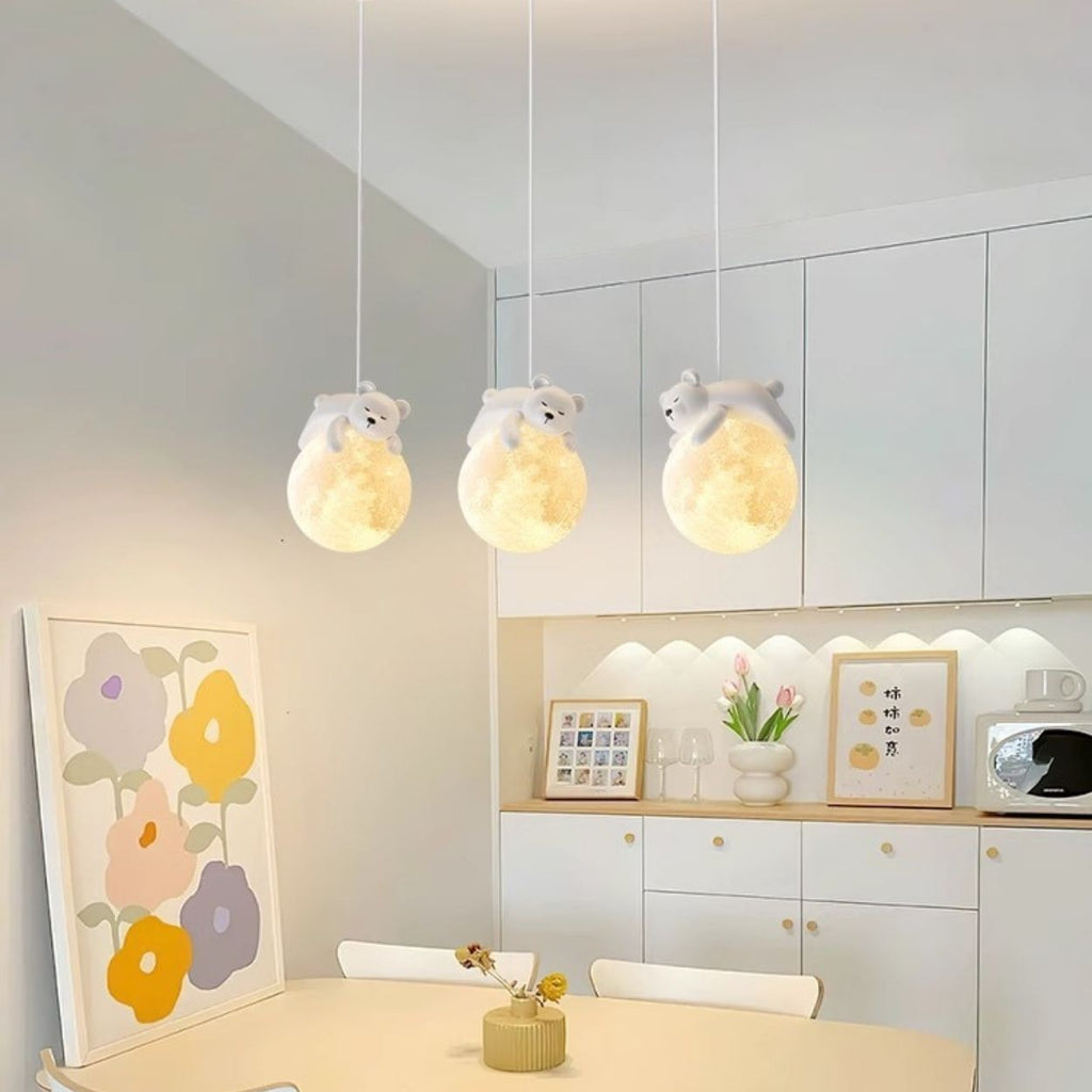 A cozy kitchen with white cabinets, colorful flower art, and three pendant lights shaped like glowing moons with small bear figurines on top, hanging above a dining table with a small vase of flowers.