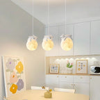 A cozy kitchen with white cabinets, colorful flower art, and three pendant lights shaped like glowing moons with small bear figurines on top, hanging above a dining table with a small vase of flowers.