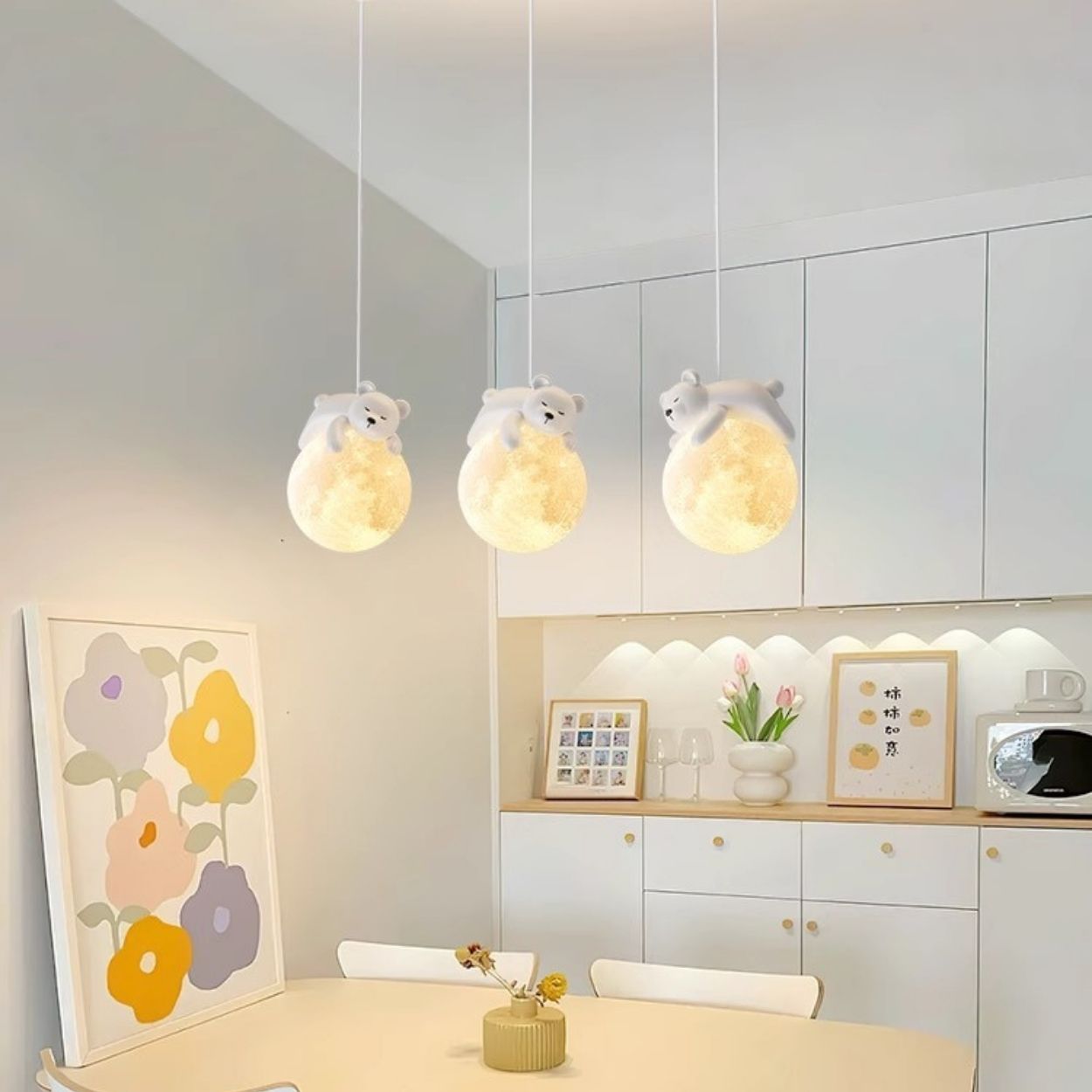 A cozy kitchen with white cabinets, colorful flower art, and three pendant lights shaped like glowing moons with small bear figurines on top, hanging above a dining table with a small vase of flowers.