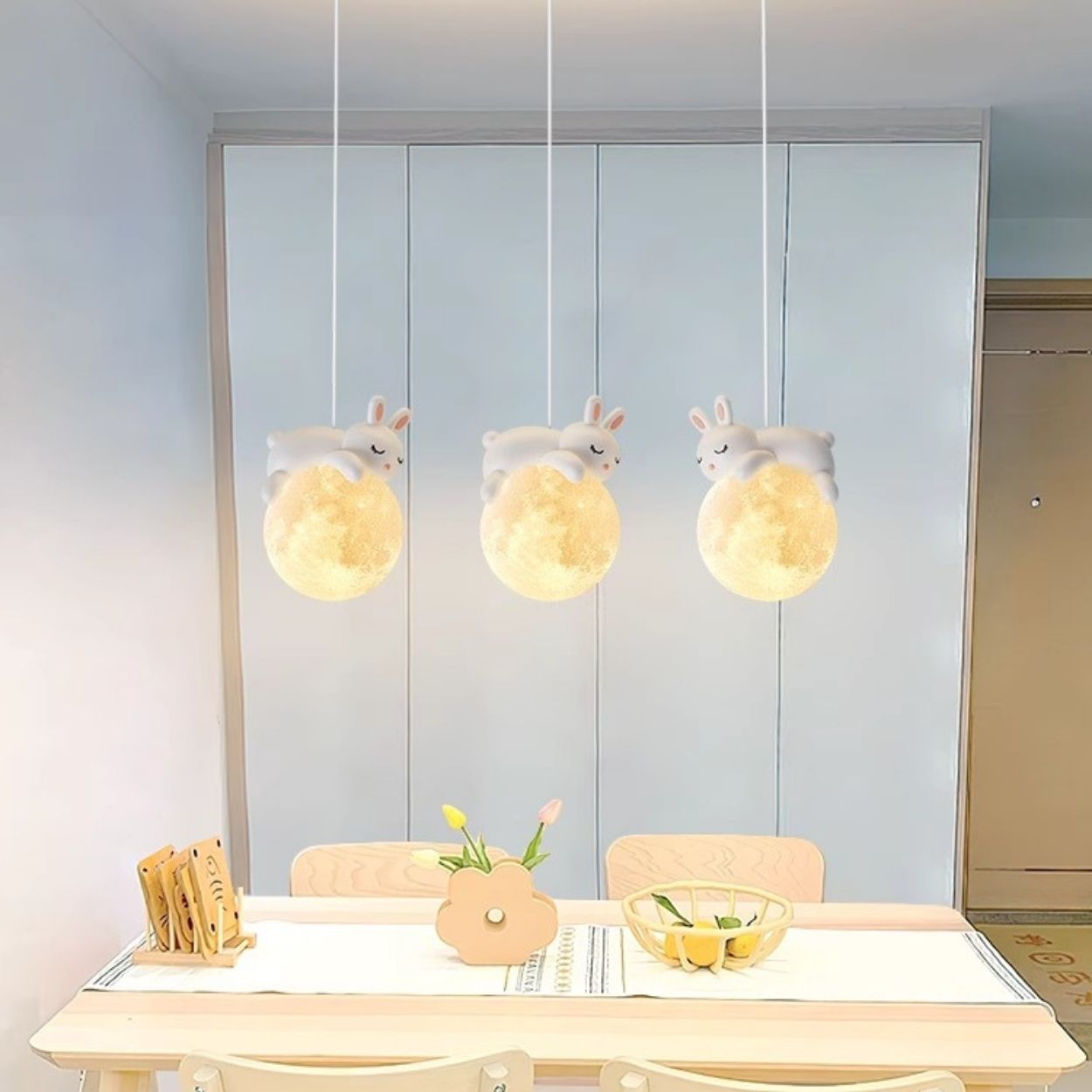 A dining area with a light wooden table, four chairs, and whimsical rabbit-shaped pendant lights sitting on glowing spheres above. The table is set with a flower vase, a bowl of fruit, and napkins for a charming touch.
