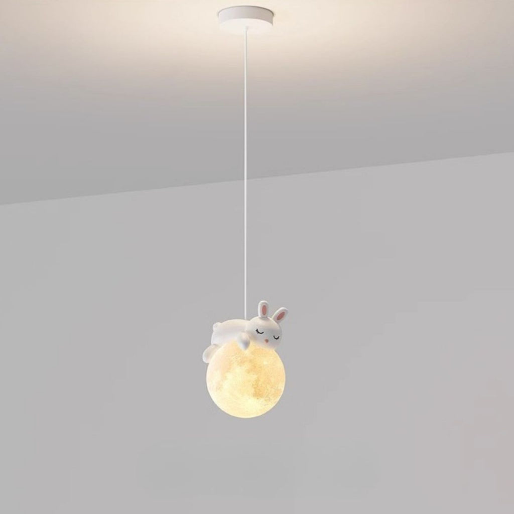 A pendant light hangs from the ceiling, featuring a glowing orb resembling the moon with a cute, white rabbit figurine lying on top. This charming pendant light is set against a plain, light gray background.