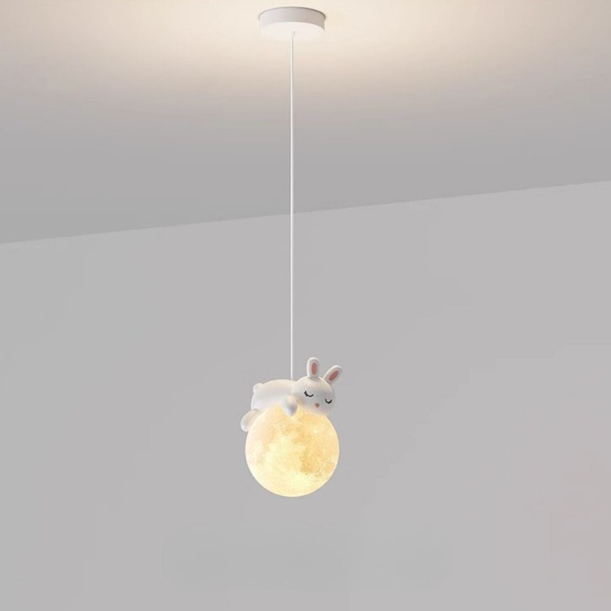 A pendant light hangs from the ceiling, featuring a glowing orb resembling the moon with a cute, white rabbit figurine lying on top. This charming pendant light is set against a plain, light gray background.