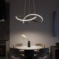 A modern dining area with a round white table, four gray chairs, and a sculptural pendant light hanging above. A black vase with decorative dried grass sits at the center of the table. The setting is dimly lit.