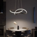A modern dining area with a round white table, four gray chairs, and a sculptural pendant light hanging above. A black vase with decorative dried grass sits at the center of the table. The setting is dimly lit.