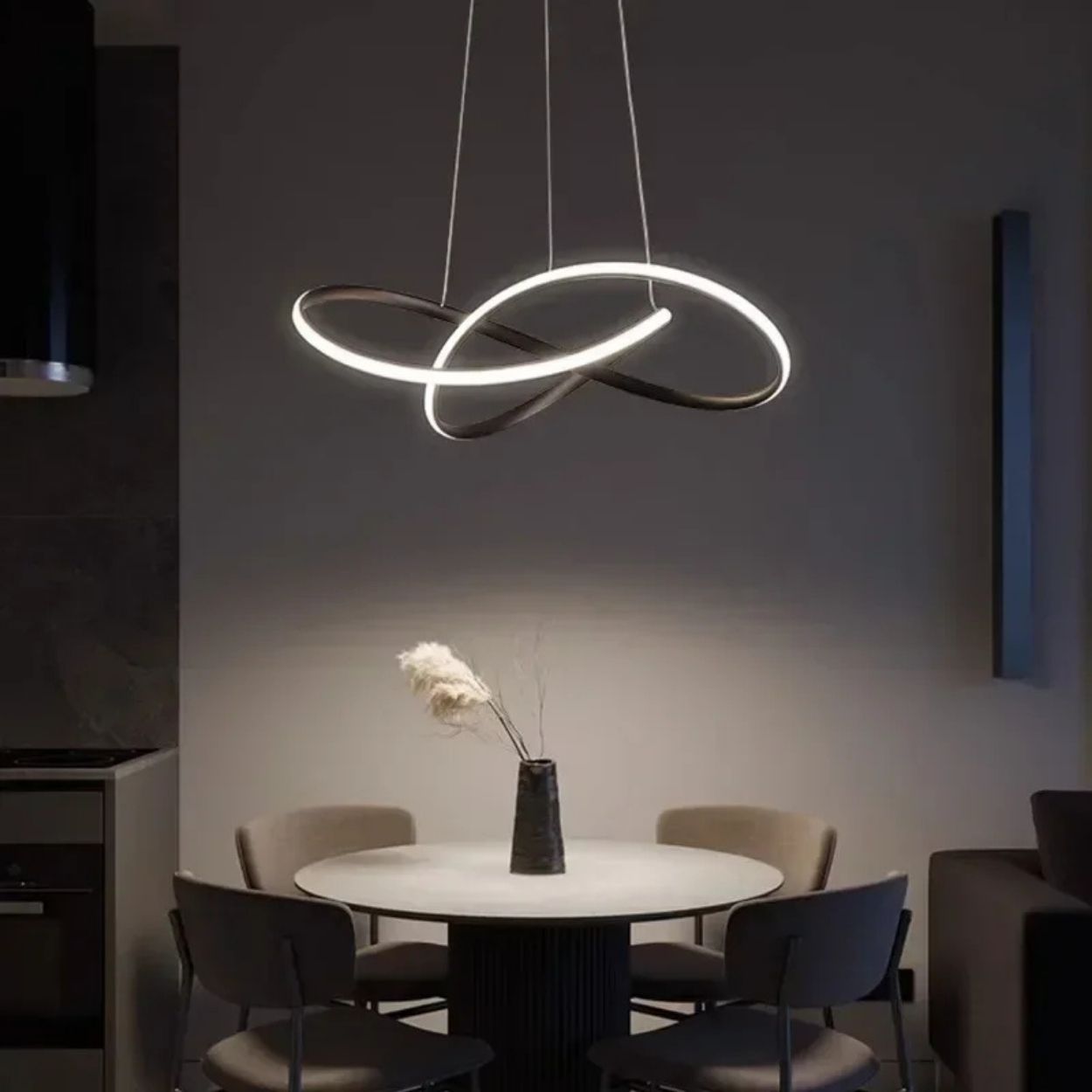 A modern dining area with a round white table, four gray chairs, and a sculptural pendant light hanging above. A black vase with decorative dried grass sits at the center of the table. The setting is dimly lit.