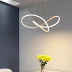 A modern, abstract pendant light with two intersecting LED loops hangs from the ceiling above a table with a vase of yellow flowers against a gray and white wall.