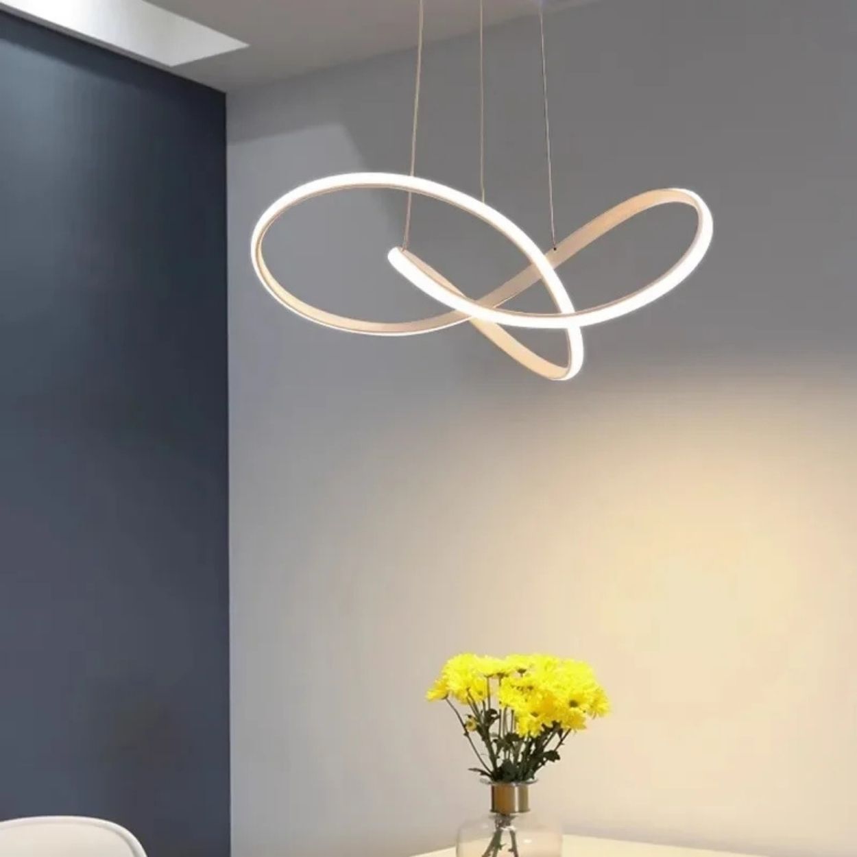 A modern, abstract pendant light with two intersecting LED loops hangs from the ceiling above a table with a vase of yellow flowers against a gray and white wall.