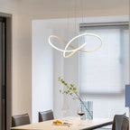 A modern dining area with a rectangular table, four chairs, wine glasses, and a decorative plant. An artistic, loop-shaped pendant light hangs above the table, while blinds cover a large window in the background.