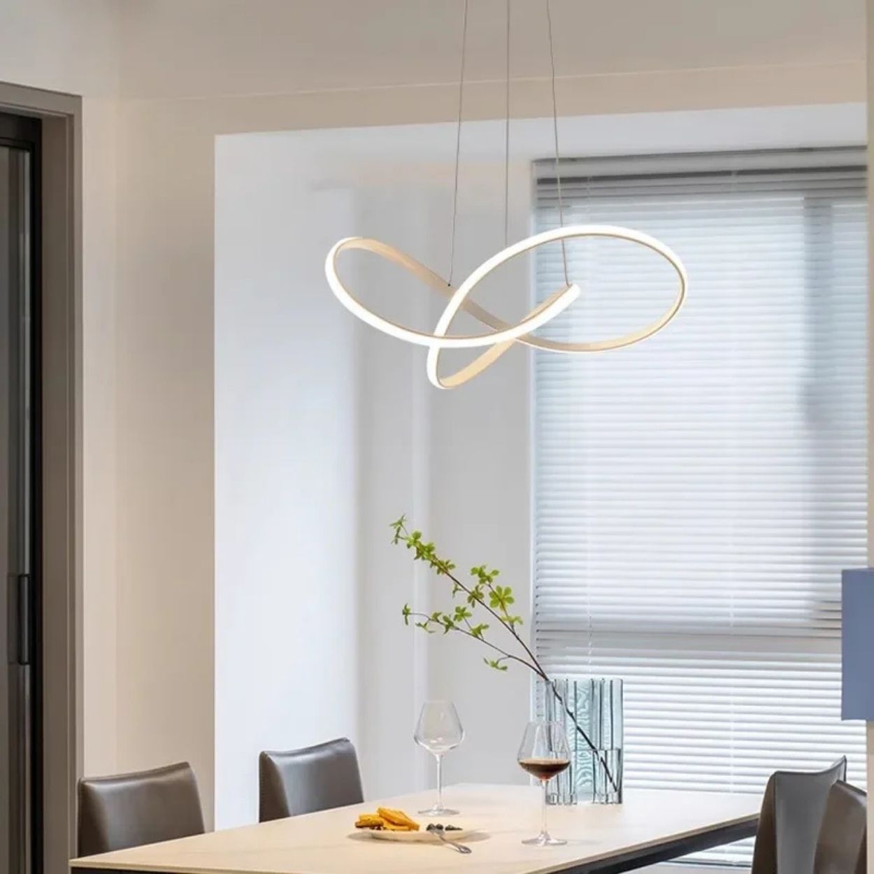 A modern dining area with a rectangular table, four chairs, wine glasses, and a decorative plant. An artistic, loop-shaped pendant light hangs above the table, while blinds cover a large window in the background.