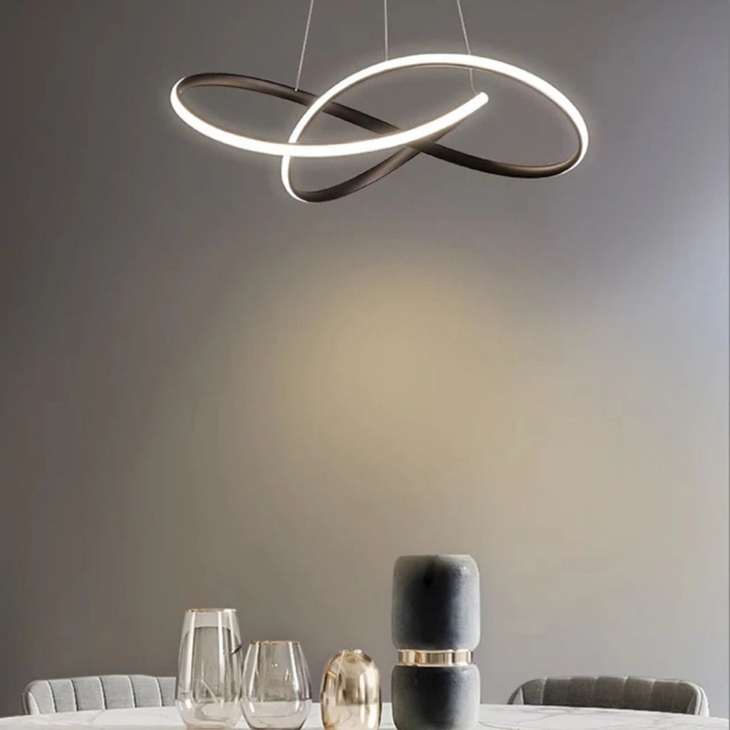 A modern dining area featuring a round table with glassware and decorative objects, illuminated by a sculptural, intertwined pendant light against a neutral gray wall.