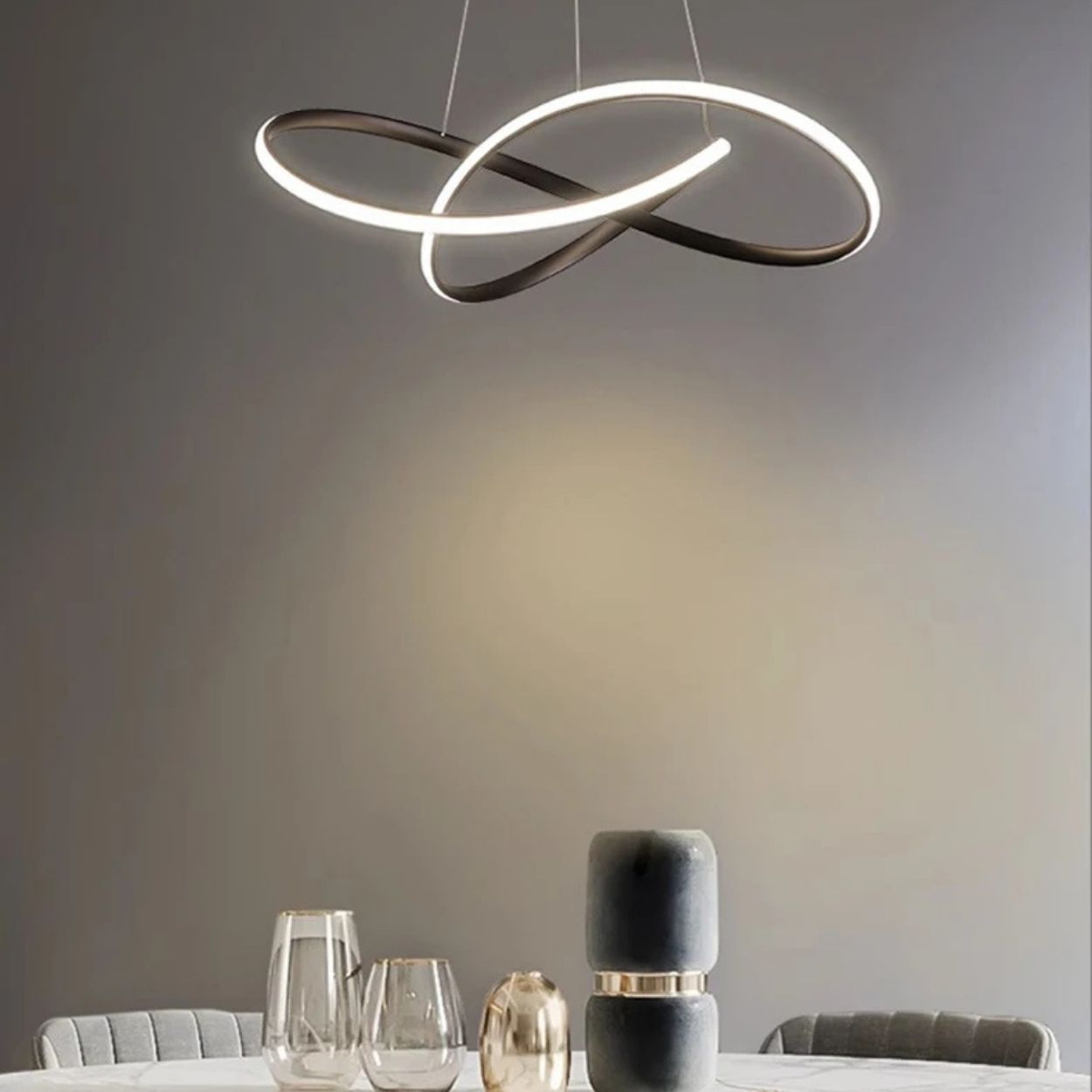 A modern dining area featuring a round table with glassware and decorative objects, illuminated by a sculptural, intertwined pendant light against a neutral gray wall.