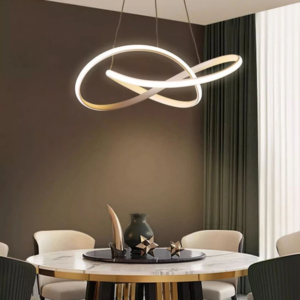 A modern dining room with a round table, black and gold chairs, and an artistic, looped LED chandelier and pendant light hanging above. The table holds decorative items, while dark curtains frame the beige walls.