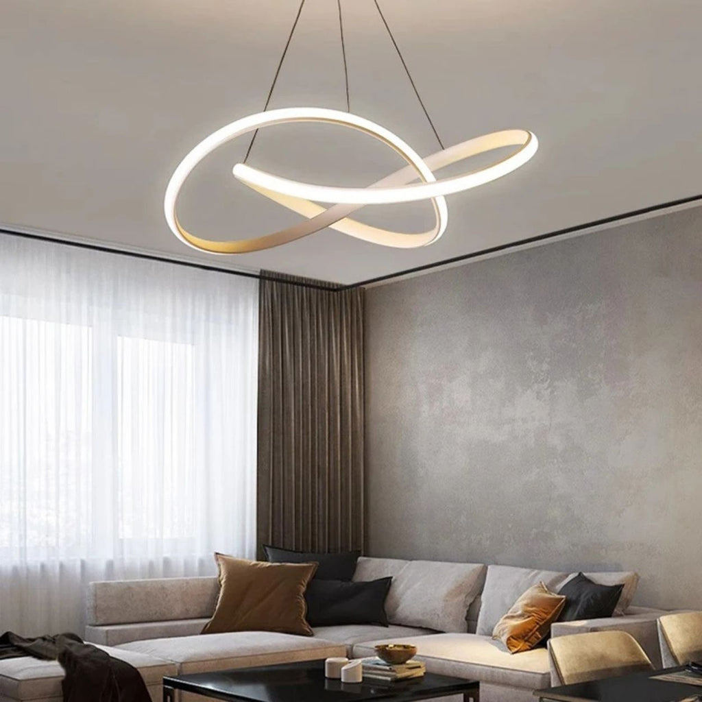 A modern living room with a sectional sofa, throw pillows, a coffee table, and a striking Pendant Light in a sculptural looping design hanging from the ceiling. Large windows are dressed with sheer and dark curtains.