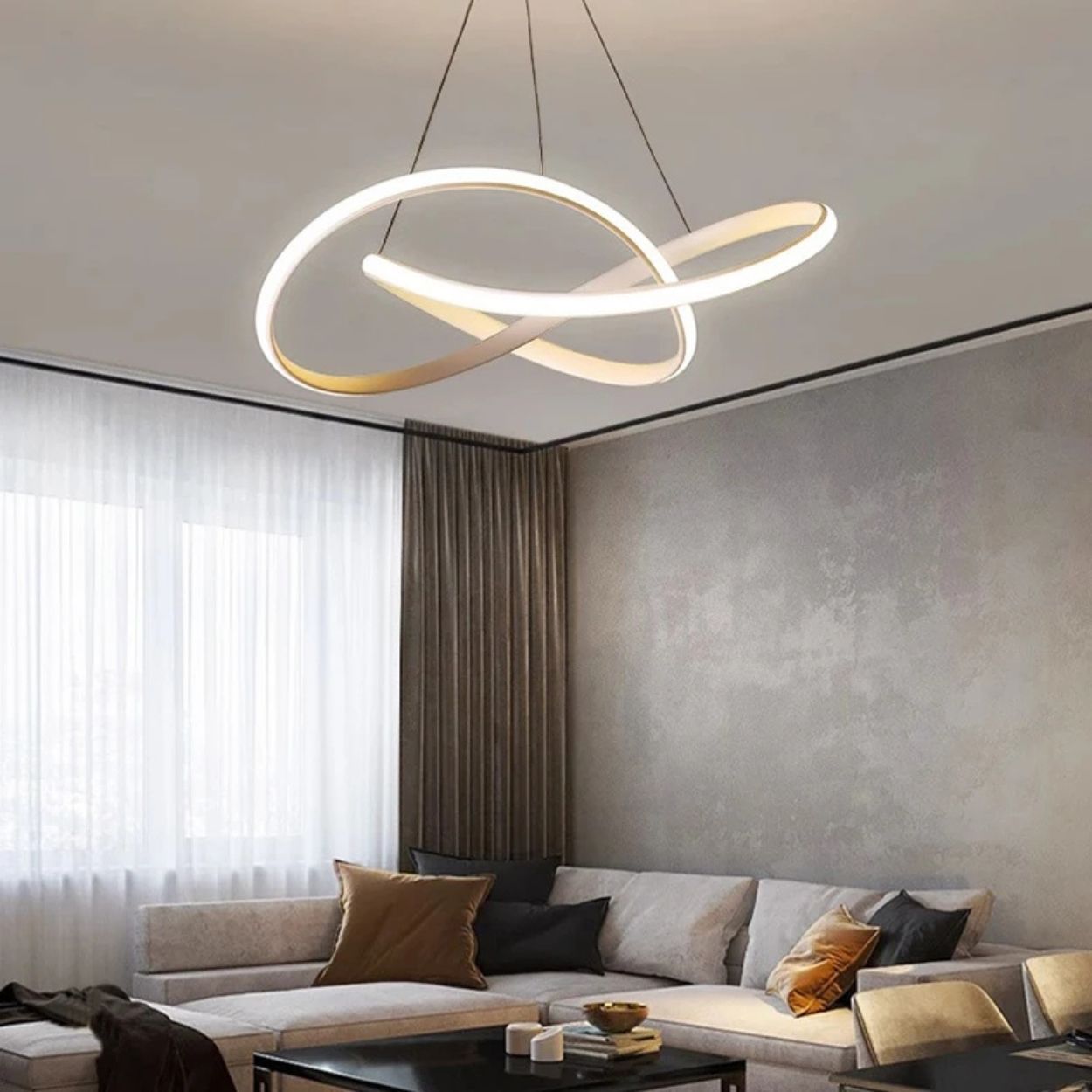 A modern living room with a sectional sofa, throw pillows, a coffee table, and a striking Pendant Light in a sculptural looping design hanging from the ceiling. Large windows are dressed with sheer and dark curtains.