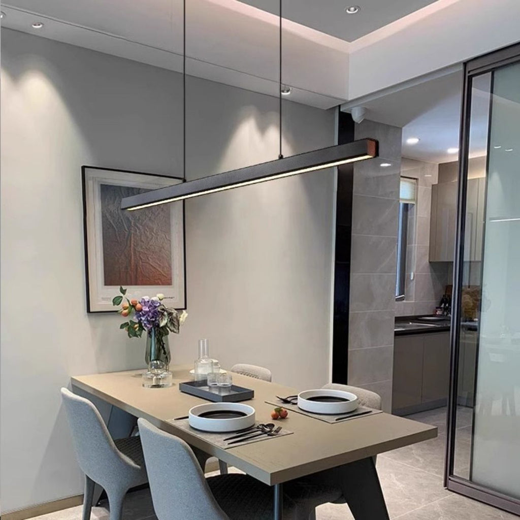 A modern dining area with a beige table set for two, featuring black and white plates, a vase of flowers, and a sleek pendant light above. The room has grey chairs and minimal, contemporary decor.