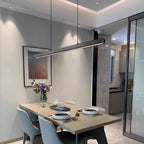 A modern dining area with a beige table set for two, featuring black and white plates, a vase of flowers, and a sleek pendant light above. The room has grey chairs and minimal, contemporary decor.