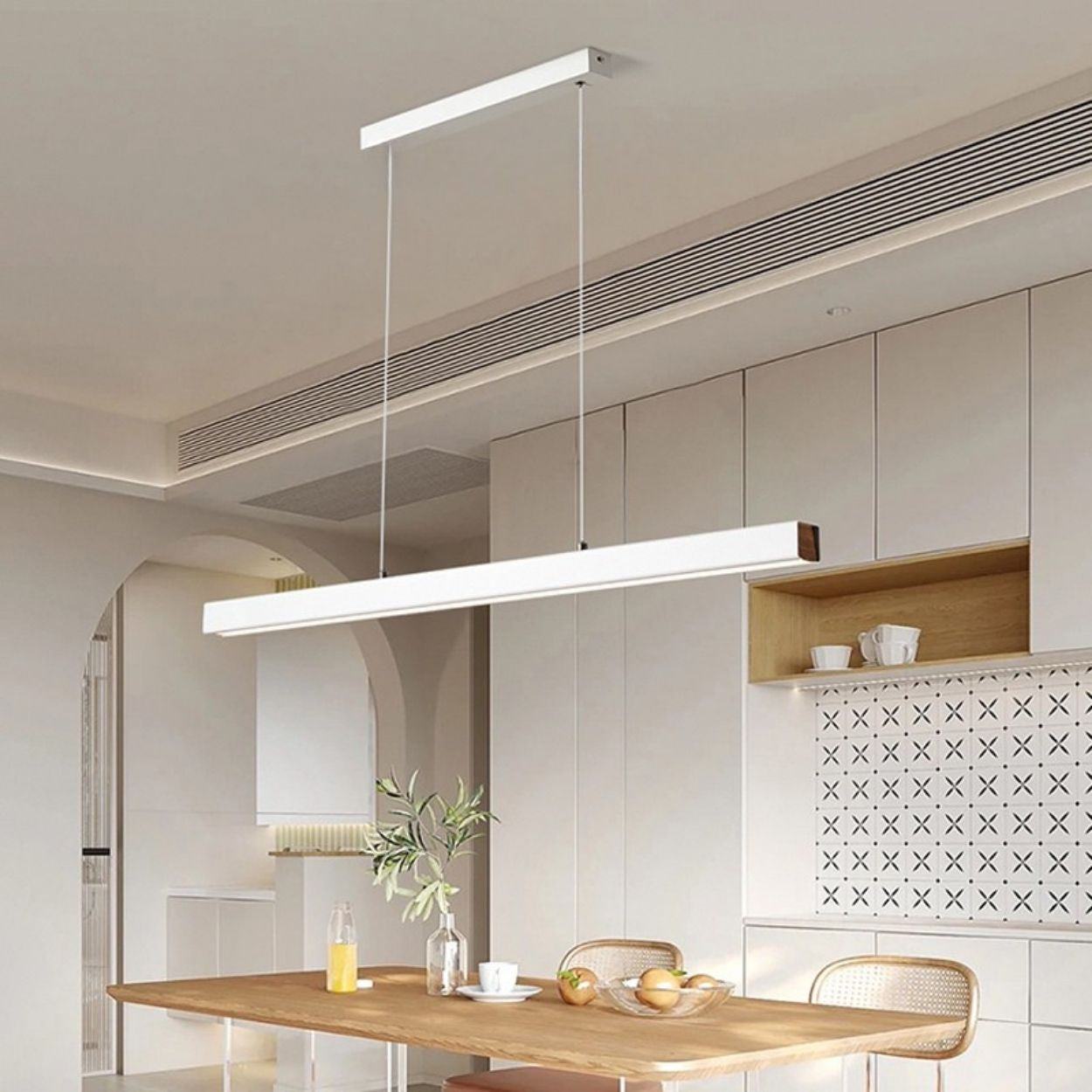 A modern, minimalist kitchen features light wood furniture and a striking rectangular pendant light above the table. Built-in cabinets with patterned backsplash tiles complete the look, with a few tableware items and a glass bottle on the table.