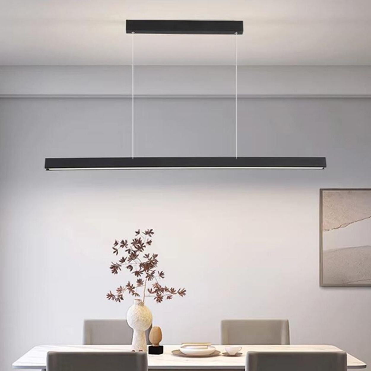 A modern dining room features a long, rectangular pendant light hanging above a white table set with a decorative vase, plates, and four chairs. A minimalist framed artwork adorns the wall.